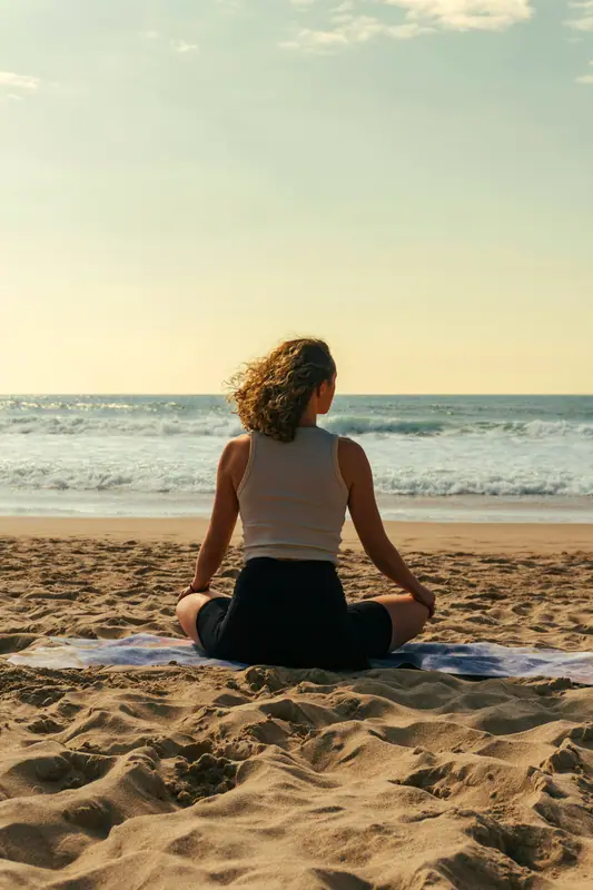 Seated meditation on a beach mat facing the ocean at golden hour, seen from behind